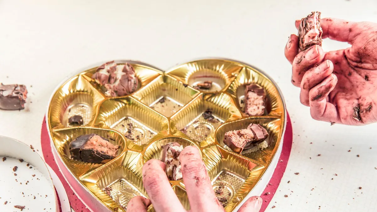 Close-up of messy hands indulging in chocolates from an empty heart-shaped box.