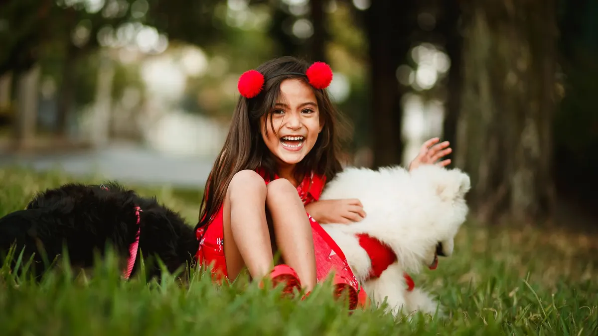 Cheerful kid in summer outfit sitting on lawn and playing with friendly dogs while looking at camera