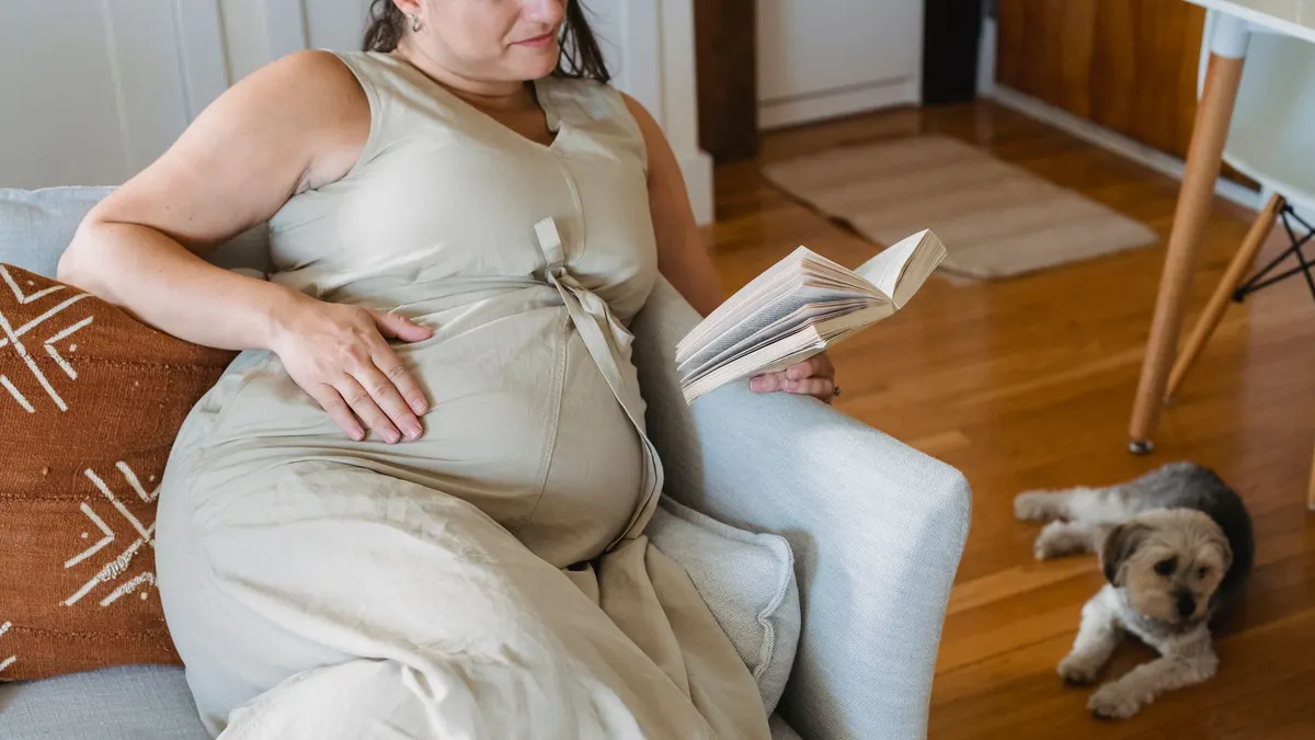 From above crop cheerful pregnant lady in casual dress sitting on comfortable couch and reading interesting book while dog lying nearby on parquet