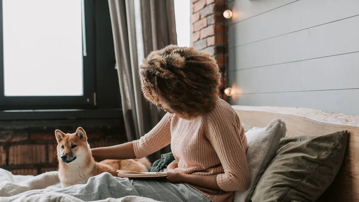 Black woman with afro hair reads on bed with her dog, cozy indoor setting.