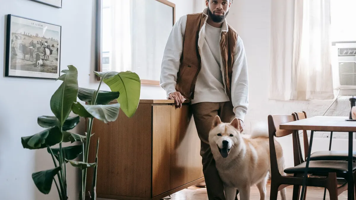 Calm young African American male owner with dreadlocks and beard standing in cozy kitchen with cute Akita Inu dog
